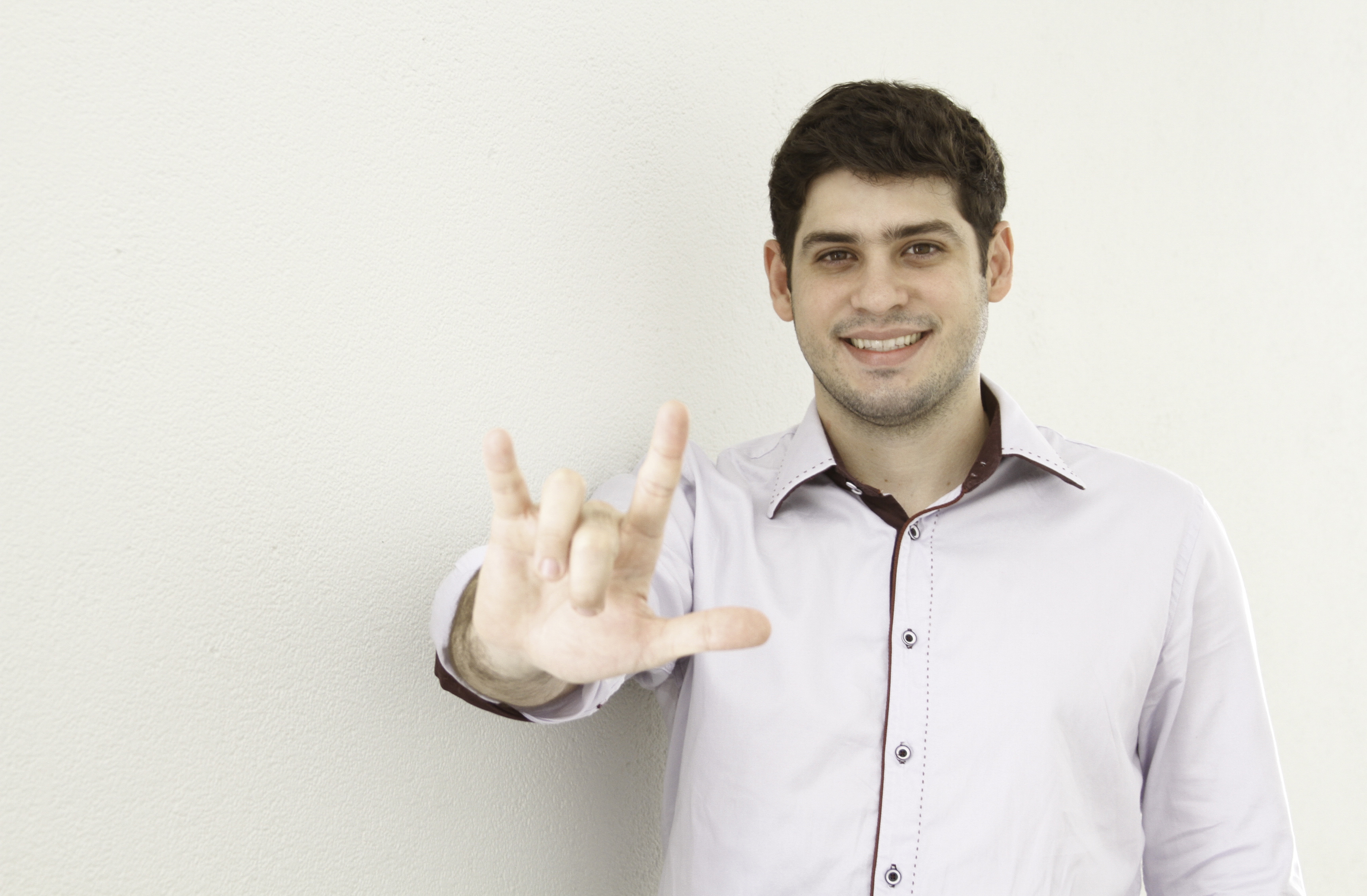 Foto de Ronaldo Tenório, CEO e cofundador da Hand Talk, de camisa branca, sorrindo e fazendo o simbolo I love you em Libras, sobre o fundo branco.