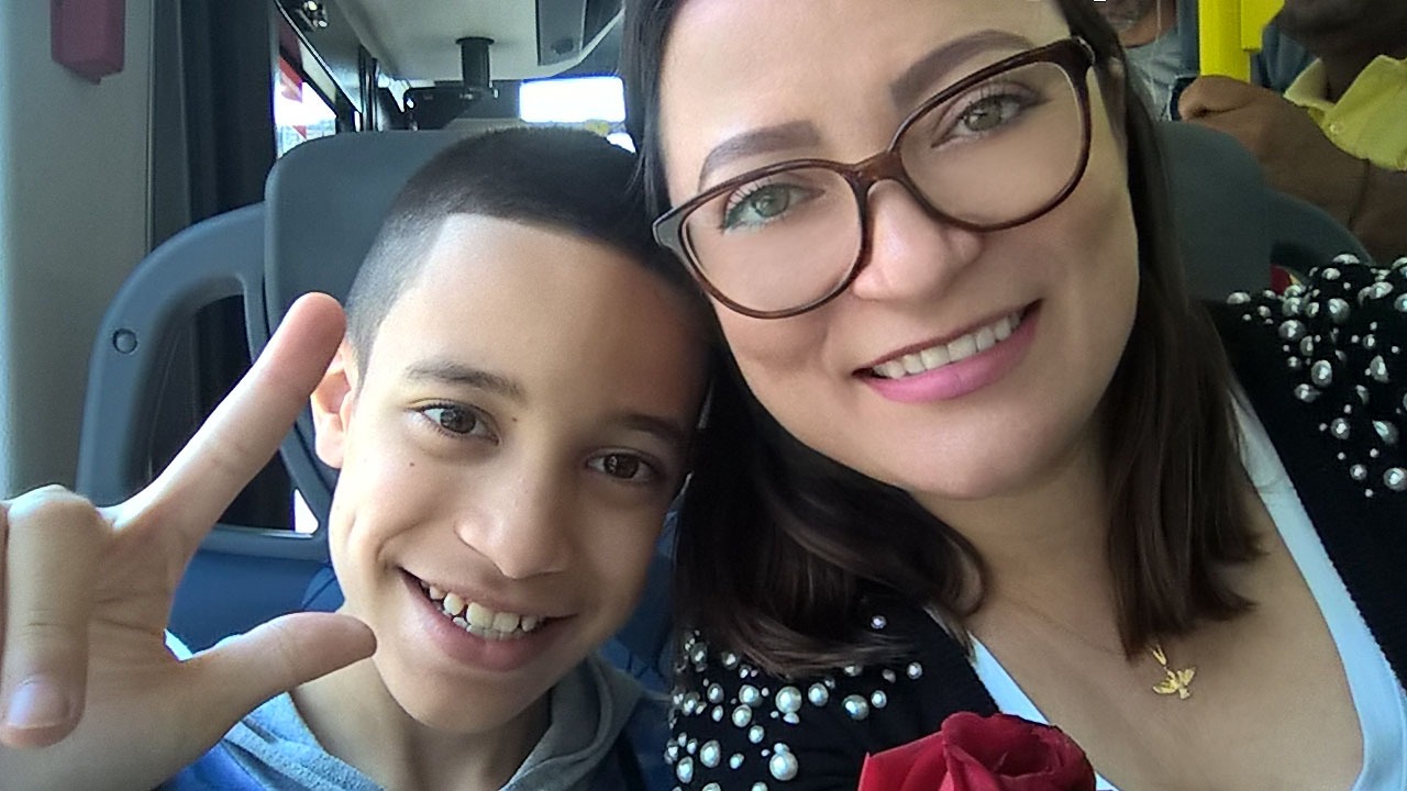 Foto da Maria com seu filho William. Maria é branca, possui olhos verdes e cabelos castanhos claros. Está usando óculo e sorrindo. William é branco, possui cabelos castanhos curtos e sorri para a foto enquanto faz o sinal de "I Love You em Libras.