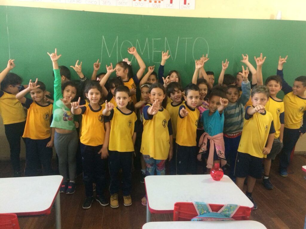 Foto de uma sala de aula com várias crianças usando o uniforme da escola, na cor amarela. Elas estão em frente a uma lousa e fazendo diferentes sinais com as mãos. 