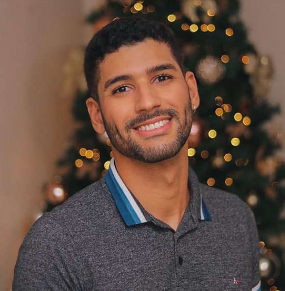 Foto de homem petro de cabelo e barbas pretos e curtos. Ele está sorrindo e vestindo camiseta polo cinza. Ao fundo uma árvore de Natal desfocada.