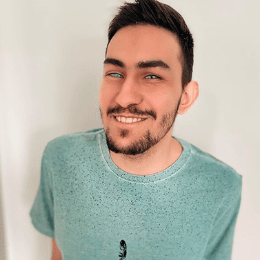 Gustavo é um homem branco, com cabelos curtos castanhos e lisos. Ele tem olhos claros, barba e bigode, e está sorrindo. Gustavo está usando uma camiseta de manga curta verde.