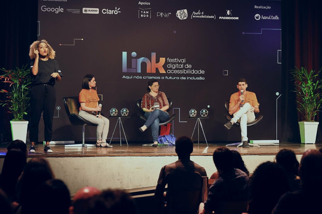 Foto demonstra pessoas em cima do palco do Link. Três pessoas estão sentadas em cadeiras, e a intérprete de libras está em pé.