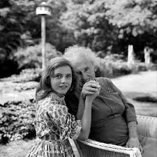 "Photo of an elderly woman (Helen Keller) holding the hand of a child (Patty Duke). The child is standing, and the elderly woman is sitting. In the background, there is a landscape. The photo is in black and white."