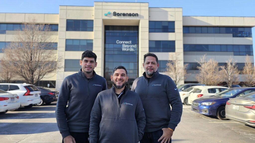 Foto dos fundadores da Hand Talk em frente ao prédio da Sorenson, nos Estados Unidos. Da esquerda para a direita, Ronaldo Tenório, Thadeu Luz e Carlos Wanderlan vestem casacos com o logo da Sorenson.