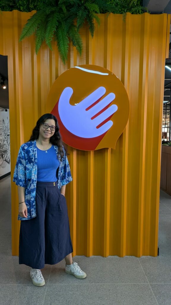 Fotografia de Betty Chu, Ela é uma mulher de cabelos castanhos, com traço orientais, que usa óculos de armação preta, vestindo roupas azuis. Ela está ao lado do logo da Hand Talk, presente em sua sede (Maceió - Alagoas - Brasil).
