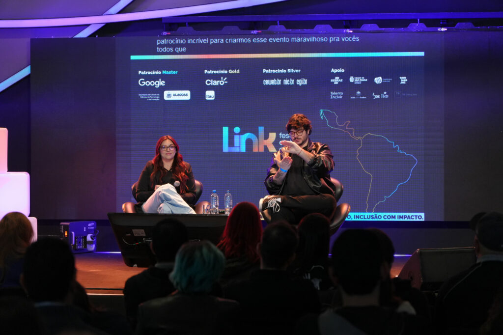 A criadora de conteúdo Letícia Fabbri e o criador e designer Roberto Castejon (Hand Talk) estão sentadas em poltronas no palco de um evento. À esquerda, uma mulher segura um microfone no colo; à direita, um homem faz gestos com as mãos enquanto fala. No fundo há um telão grande com logotipos de patrocinadores e o nome do evento “Link Festival”. Algumas pessoas do público aparecem em primeiro plano, de costas.