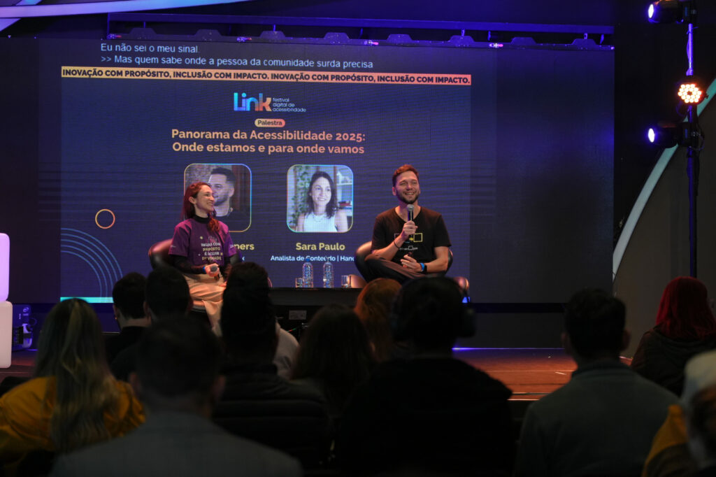 Sara Paulo (Hand Talk) e Felipe Schepers (Opinion Box) estão sentados em poltronas em um palco, sorrindo e segurando microfones. Atrás deles, um telão mostra o título da palestra ‘Panorama da Acessibilidade 2025: Onde estamos e para onde vamos’, com fotos e nomes dos palestrantes. Pessoas da plateia aparecem de costas, assistindo.