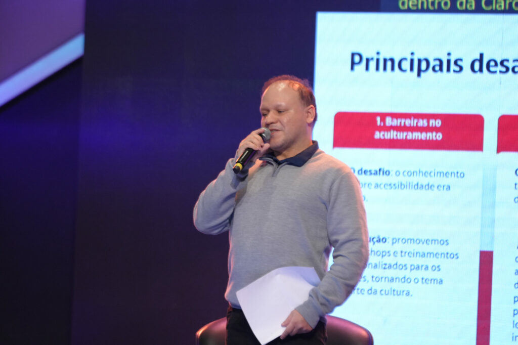 Éber Anacleto veste suéter cinza claro e segura um microfone em uma mão e uma folha na outra. Fala sorridente em frente a um telão com o título "Principais desafios" e tópicos em vermelho.