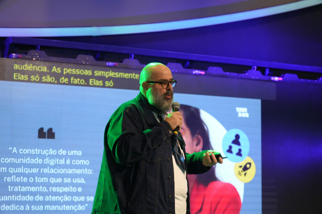 Vitor Bastos (Tambor) é barbudo e de óculos fala ao microfone em frente a um telão com texto e imagens sobre construção de comunidade digital. Ele usa jaqueta jeans escura.