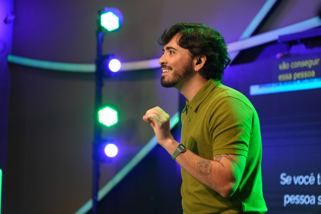 Gabriel Isaac usa barba curta, cabelos escuros e camisa polo verde fala no palco. Ele sorri e gesticula, iluminado por refletores verdes e roxos