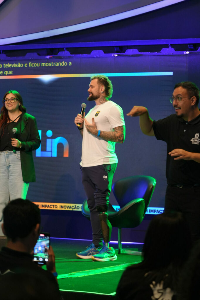 No palco, Letícia Fabbri usa óculos e blazer preto está ao lado de Vinni Rodrigues, tatuado que usa prótese na perna direita e camiseta branca, falando ao microfone. À direita, um intérprete de Libras traduz sua fala para o público.