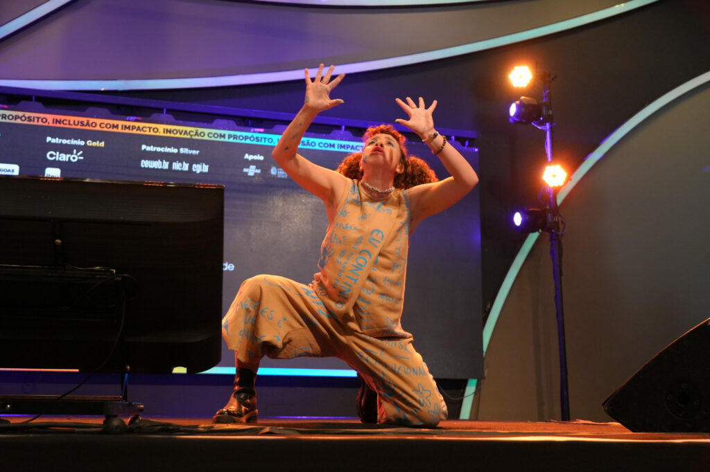Catharine Moreira está no palco, ajoelhada com os braços erguidos para cima, em uma expressão artística e performática. Usa um macacão bege estampado com palavras azuis e botas pretas.
