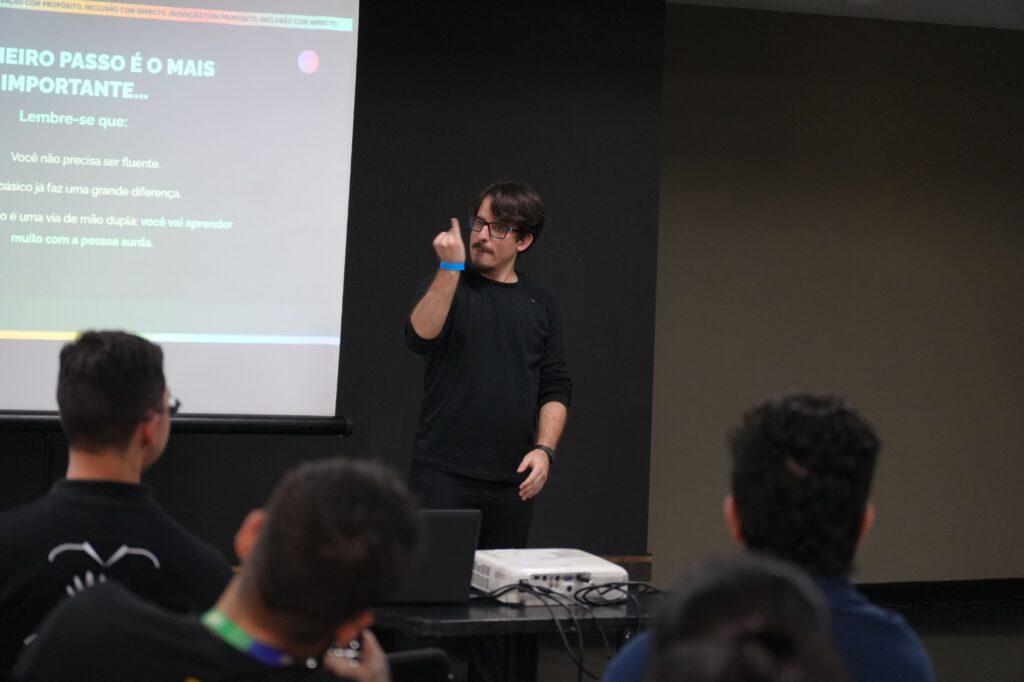 Darley Goulart (Hand Talk) usa óculos e barba curta veste camiseta preta e faz sinais em Libras em frente a uma tela de projeção com o título: “O primeiro passo é o mais importante...”. A plateia aparece em primeiro plano.
