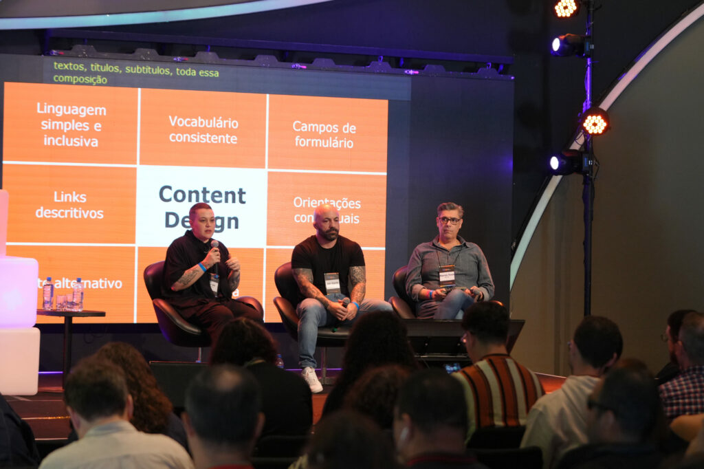 Luiza Rolim, Vitor Dantas e Maurício Pereiro (Itaú) estão sentados em poltronas pretas no palco discutem em frente a um telão laranja que exibe palavras como “Linguagem simples e inclusiva”, “Vocabulário consistente” e “Content Design”. O público assiste em primeiro plano.