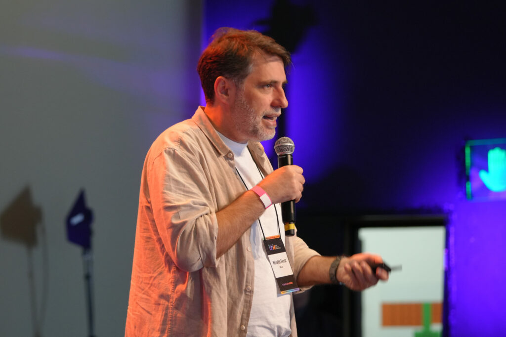 Reinaldo Ferraz (Ceweb.br) usa barba grisalha e camisa clara fala ao microfone. Ele segura um controle remoto na outra mão, em frente ao público.