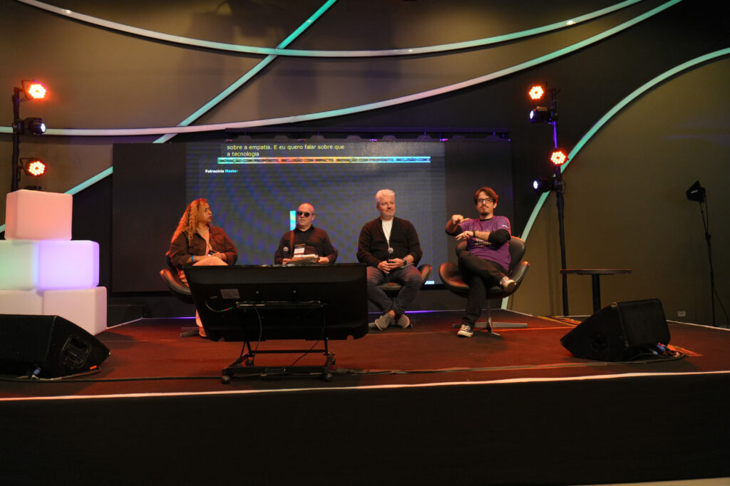 Nayara Silva, Sergio Faria (Accenture), Ricardo Wagner (Start.se) e Darley Goulart (Hand Talk) estão sentados em poltronas no palco participam de uma mesa redonda. À esquerda, uma mulher fala ao microfone; ao centro, dois homens escutam; à direita, um homem com camiseta roxa faz gestos com as mãos.