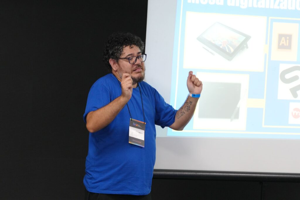 Lucas Ramon (Tikinho) usa óculos, cabelos cacheados curtos e barba rala apresenta diante de um telão com imagens de softwares de design e ilustração. Ele usa camiseta azul e crachá.