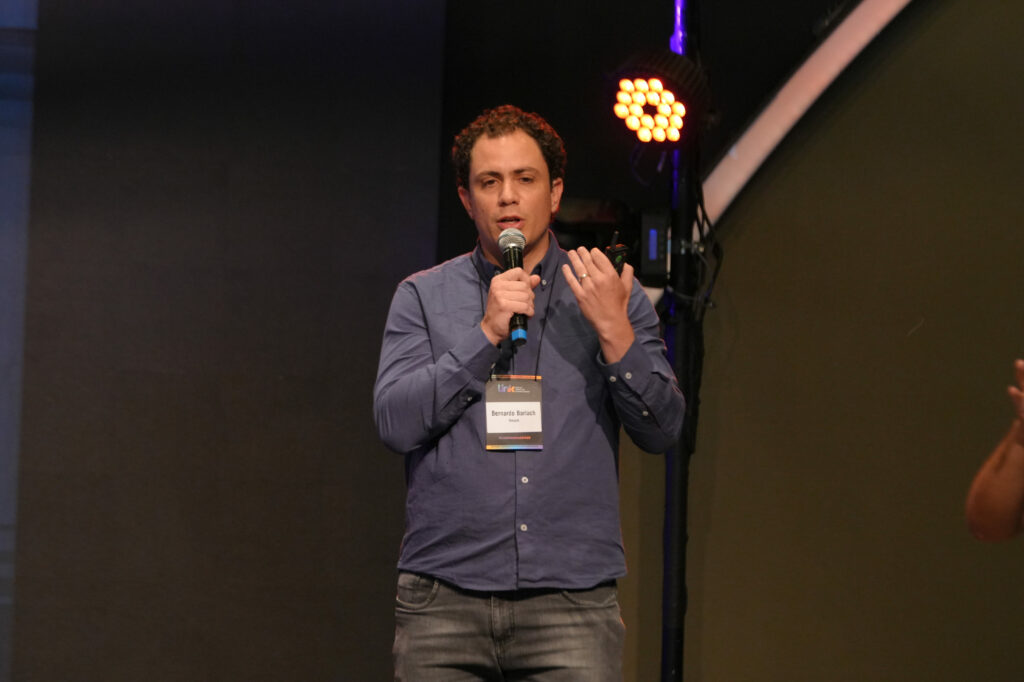 Bernardo Barlach (Google) usa camisa azul fala ao microfone, segurando um controle remoto. Ele usa crachá do evento e está iluminado por refletores roxos.