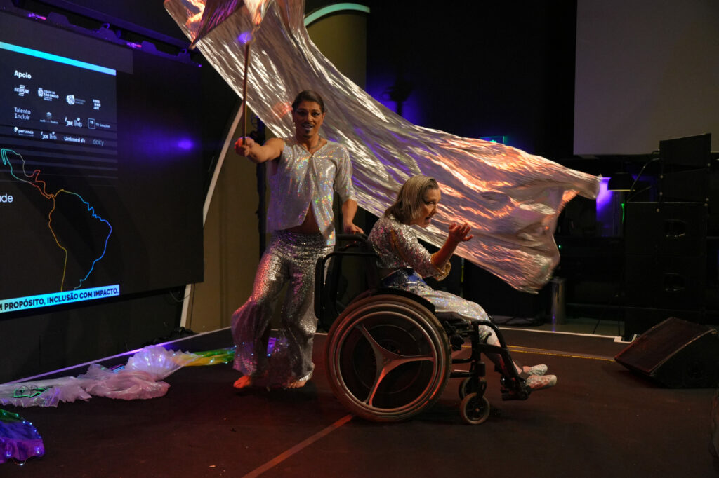 Apresentação Cia De Dança De Rodas Para o Ar: Dois artistas em apresentação no palco: à frente, Leda está em uma cadeira de rodas gesticula com intensidade; atrás dela, Clayton Brasil com roupas prateadas e capa brilhante segura um tecido longo e cintilante que se movimenta no ar.