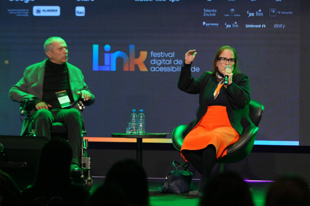 Deborah Kelly Affonso (MPT- SP) e Cid Torquato (ICOM) participam de um painel no palco. À esquerda, um homem em cadeira de rodas; à direita, uma mulher de roupa laranja vibrante fala ao microfone gesticulando com a mão. O telão atrás exibe a identidade visual do evento: “Link – festival digital de acessibilidade”.