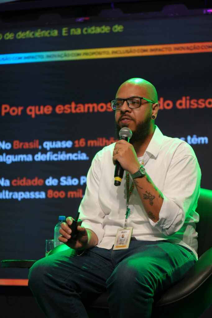 Danilo Souza (SMP - SP) usa camisa branca e óculos fala ao microfone durante sua apresentação. Ele segura um controle de slides e está sentado em uma poltrona no palco, com fundo escuro exibindo legendas em destaque.