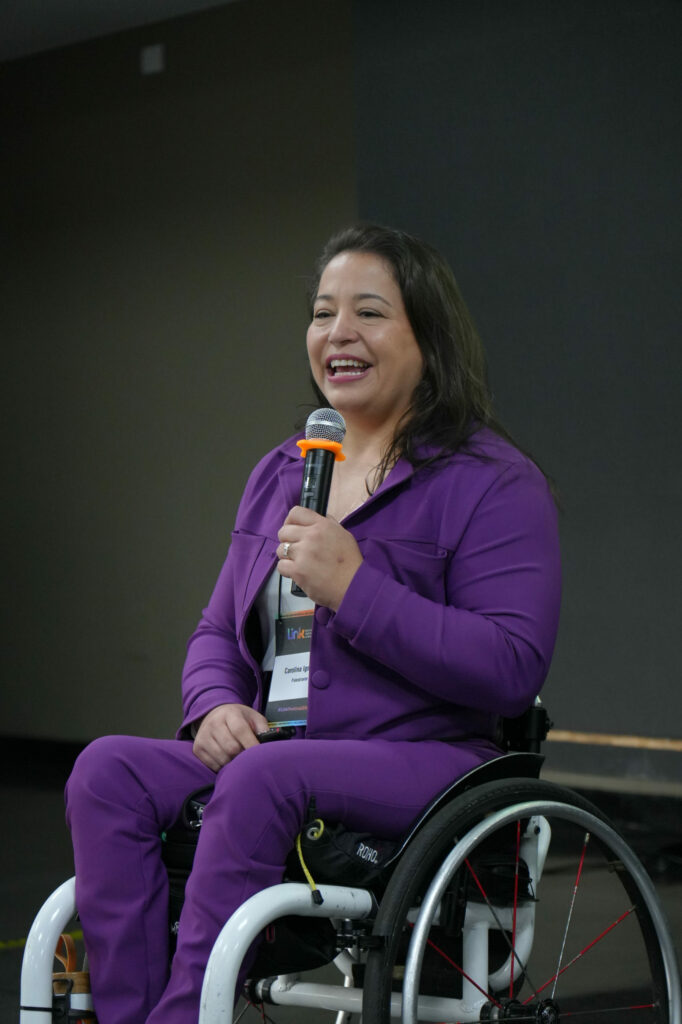 Carolina Ignarra (Talento Incluir) usa cadeira de rodas fala sorridente ao microfone. Veste um conjunto roxo e crachá do evento.