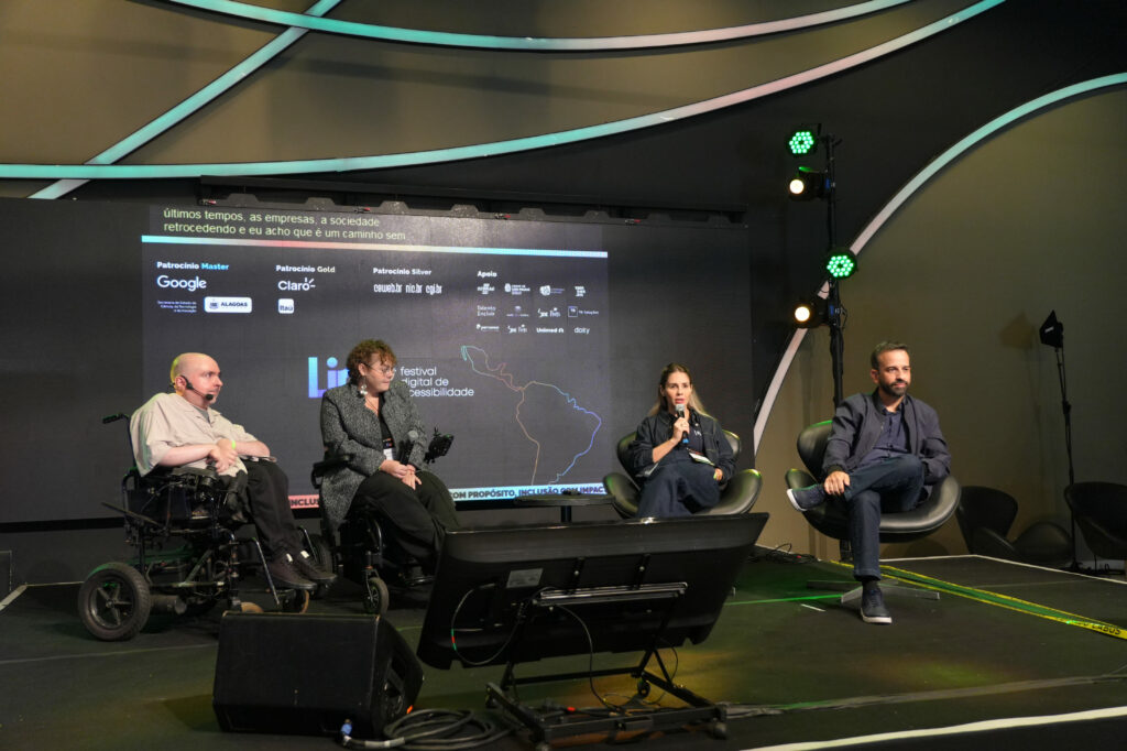 Os palestrantes Rogerio Miranda (Mosaic), Julia Piccolomini (Parada do Orgulho PCD), Manuela Alves (B3) e Tiago Bezerra (Hand Talk) participam de um painel em um palco. Duas delas estão em cadeiras de rodas, uma mulher segura o microfone enquanto fala, e um homem sentado em uma poltrona escuta. Atrás, um telão exibe logotipos de patrocinadores e o título ‘Festival Digital de Acessibilidade’.”