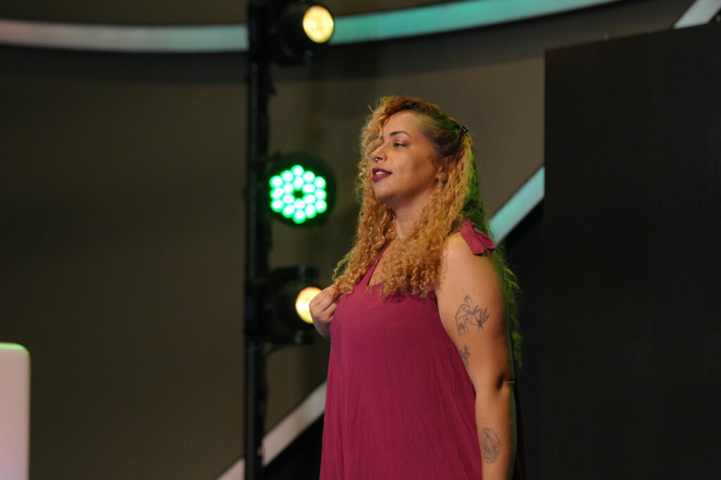 Nayara Silva usa cabelos cacheados loiros e longos fala no palco, vestida com um vestido vinho sem mangas. Ela tem tatuagens visíveis no braço e ombro.