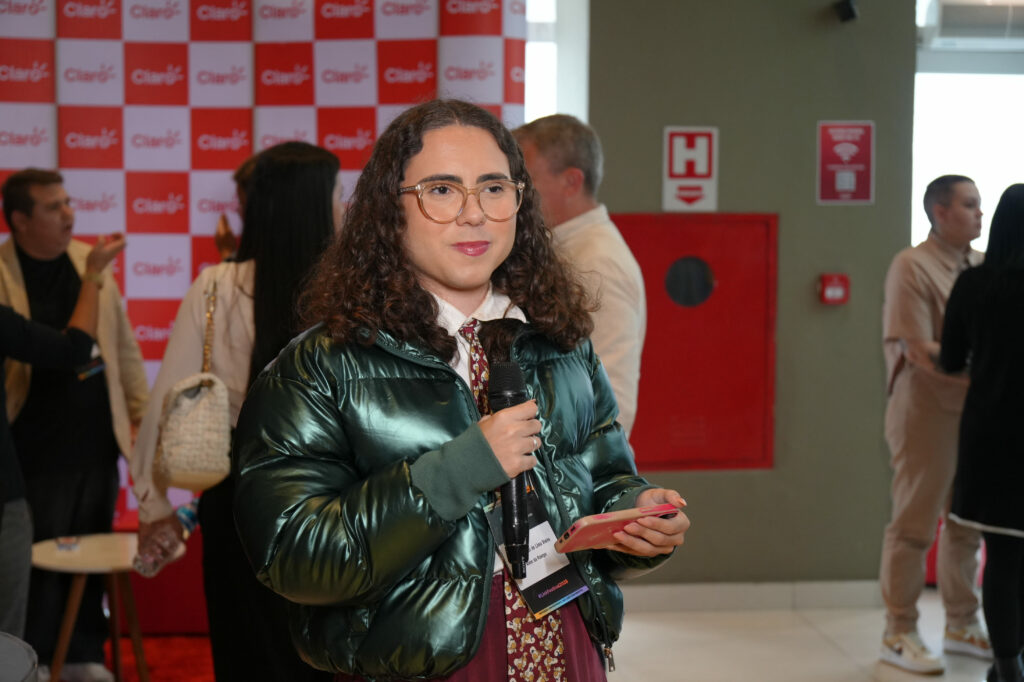 Eduardo Victor, sorridente, segura um microfone e celular, vestindo casaco verde metálico e óculos. Está em um espaço de convivência com outras pessoas ao fundo, em frente a um painel com o logo da Claro.