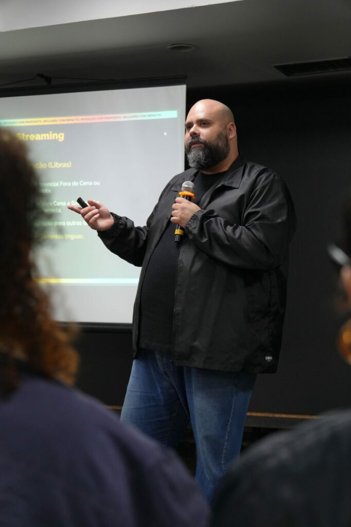 Flávio Bona (Catuaí Conteúdos) veste jaqueta preta e jeans, fala ao microfone enquanto segura um controle. Ele apresenta diante de uma tela com tópicos sobre acessibilidade em streaming.