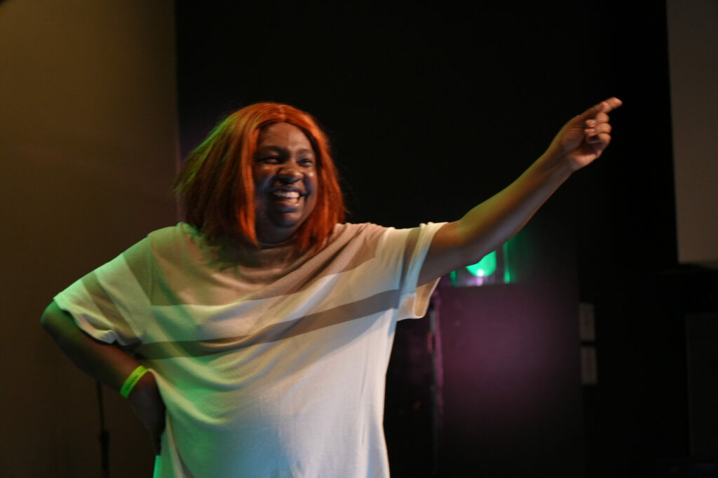 Marcos Vinni usa peruca ruiva, sorridente e descontraída, participa de uma performance no palco. Aponta para frente com expressão alegre.