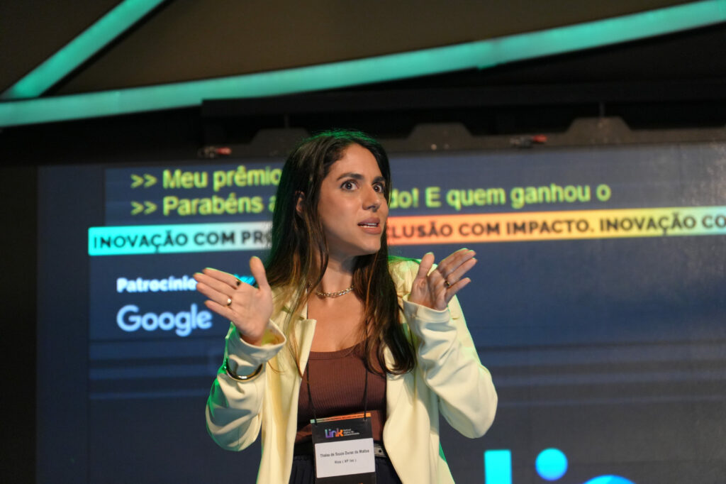 Thaisa Durso (Rico / XP) no palco fala com expressão enfática, usando crachá e blazer claro. Ao fundo, o telão exibe legendas com frases sobre inovação e inclusão.