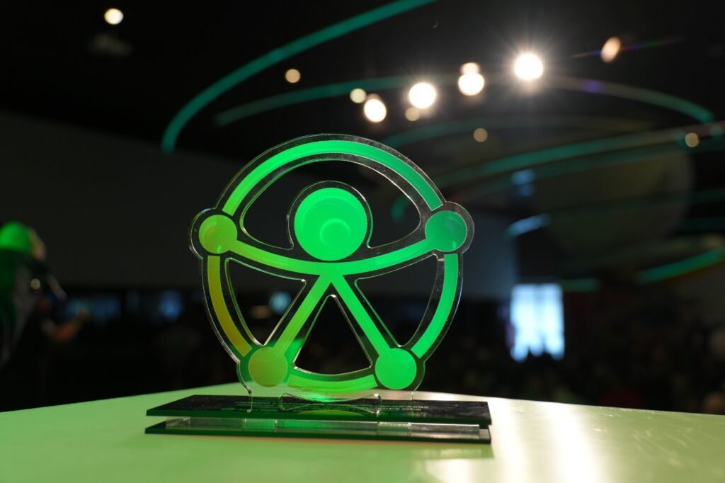 Troféu de acrílico em formato do símbolo de acessibilidade universal, iluminado em verde, sobre uma mesa. Ao fundo, o auditório aparece desfocado com luzes e detalhes arquitetônicos modernos.
