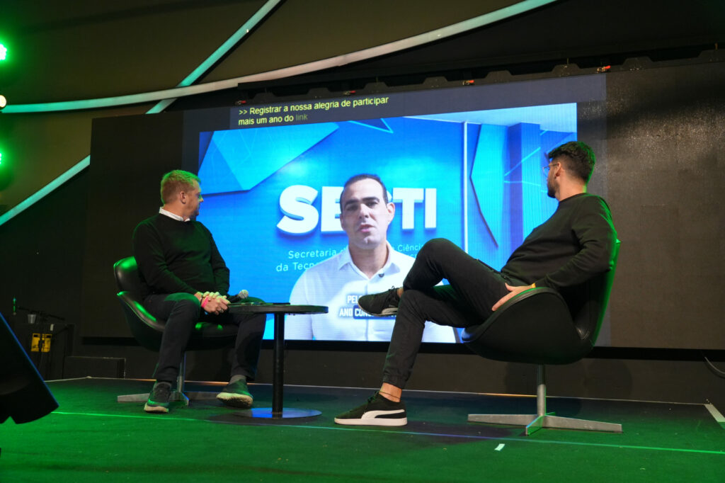 Representantes do SEBRAE, e Hand Talk estão sentados no palco, olhando para um grande telão que exibe a fala gravada do representante do SECTI, em uma transmissão remota. O telão mostra o logotipo da SECTI (Secretaria de Estado da Ciência, da Tecnologia e da Inovação).