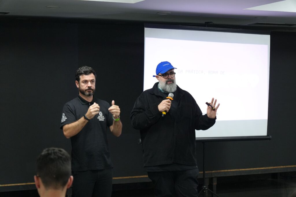Vitor Bastos (Tambor.biz) se apresenta junto com intérprete. À esquerda, um intérprete de Libras com camiseta preta e logo do Governo de São Paulo. À direita, um homem de barba grisalha, óculos e boné azul fala ao microfone gesticulando.