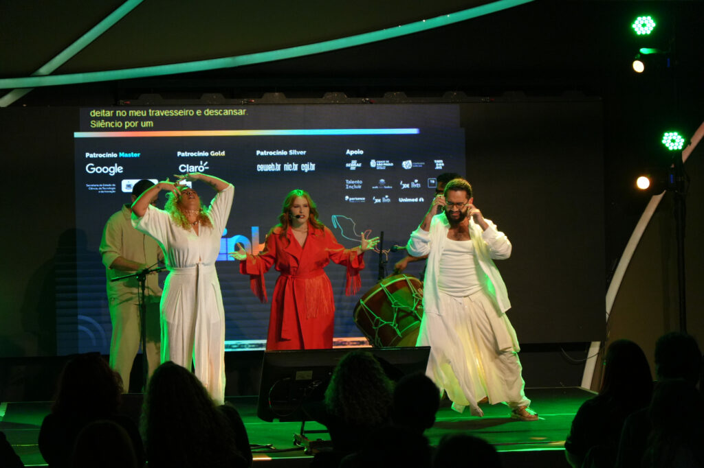 Apresentação musical de Luiza Caspary no palco com três artistas principais: uma pessoa de roupa branca ergue os braços para cima, outra de vermelho canta ao microfone e uma terceira de branco dança. Ao fundo, músicos acompanham a performance.