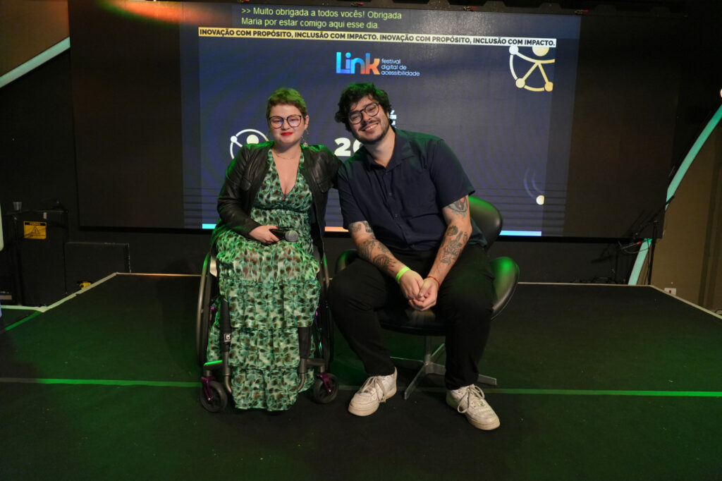 A criadora Maria Paula e o criador e designer Roberto Castejon (Hand Talk) estão sentados lado a lado no palco de um evento. Ambos sorriem para a câmera. Ela segura um microfone e veste um vestido estampado verde; ele veste camisa escura e tênis brancos. Ao fundo, há um telão com o nome “Link Festival Digital de Acessibilidade” e a frase “Inovação com propósito, inclusão com impacto”.