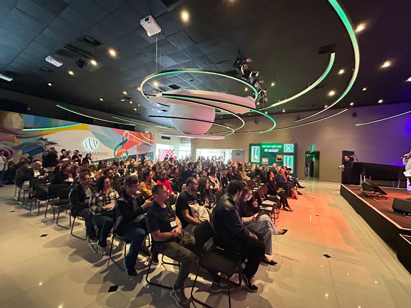 Auditório amplo e moderno, com teto preto e iluminação em linhas curvas verdes, repleto de pessoas sentadas assistindo a uma apresentação no palco à direita. A plateia é diversa, composta por homens e mulheres de diferentes idades. No fundo, há murais coloridos e um painel iluminado com símbolos de acessibilidade.