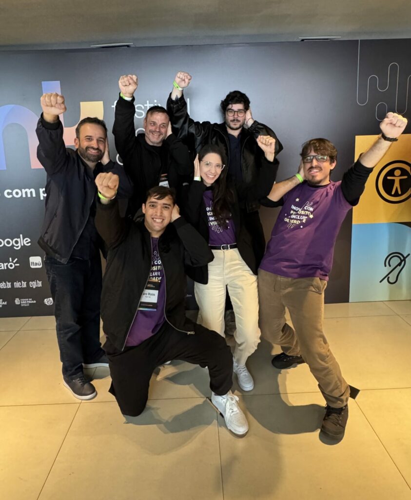 Seis HandTalkers surdos estão sorrindo enquanto fazem a pose do Deaf Power: uma mão posicionada na sua própria orelha e a outra mão levantada, conectando a surdez com uma posição de poder.