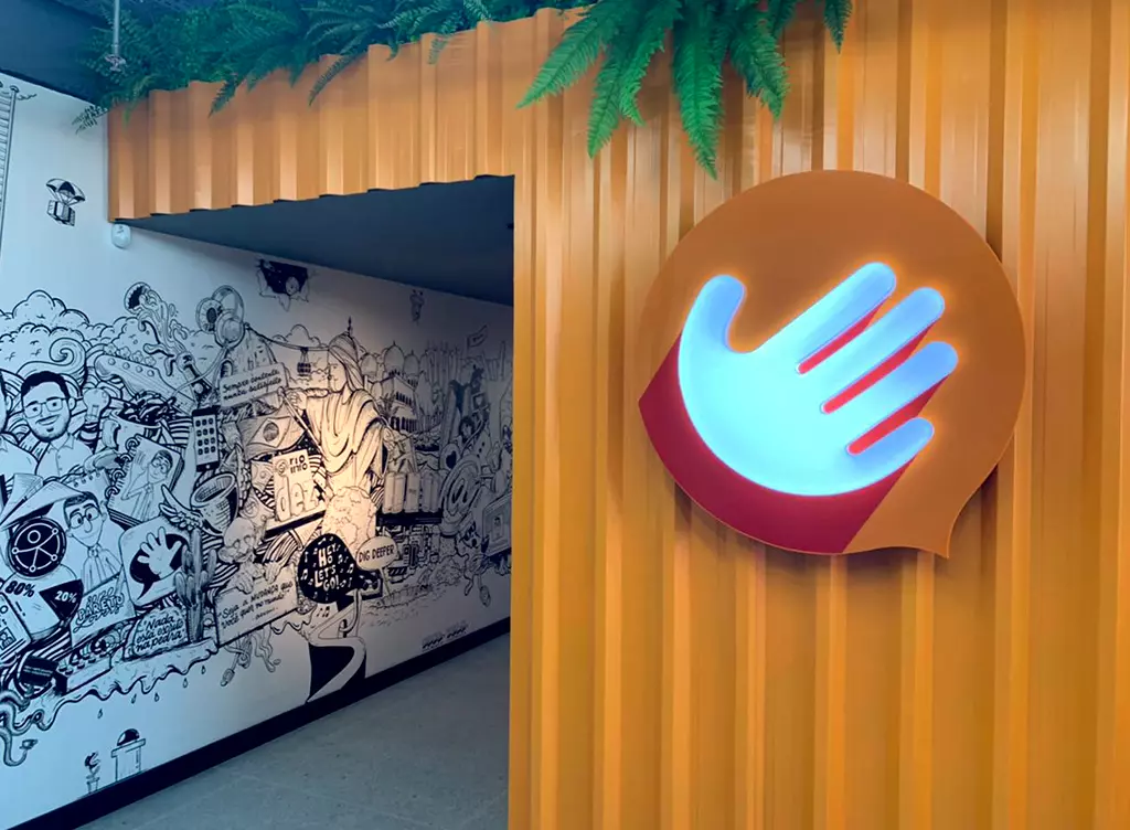 Picture of the entrance of the new Hand Talk headquarter. There is a white wall with multiple drawings on it on the left, and an orange wall on the right with the Hand Talk logo on it.