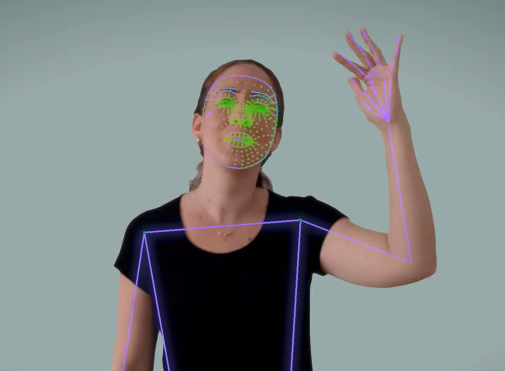 Picture of a woman signaling in Sign Language. There are purple and green lines on top of her, from a movement recognition technology. She is in front of a white wall.