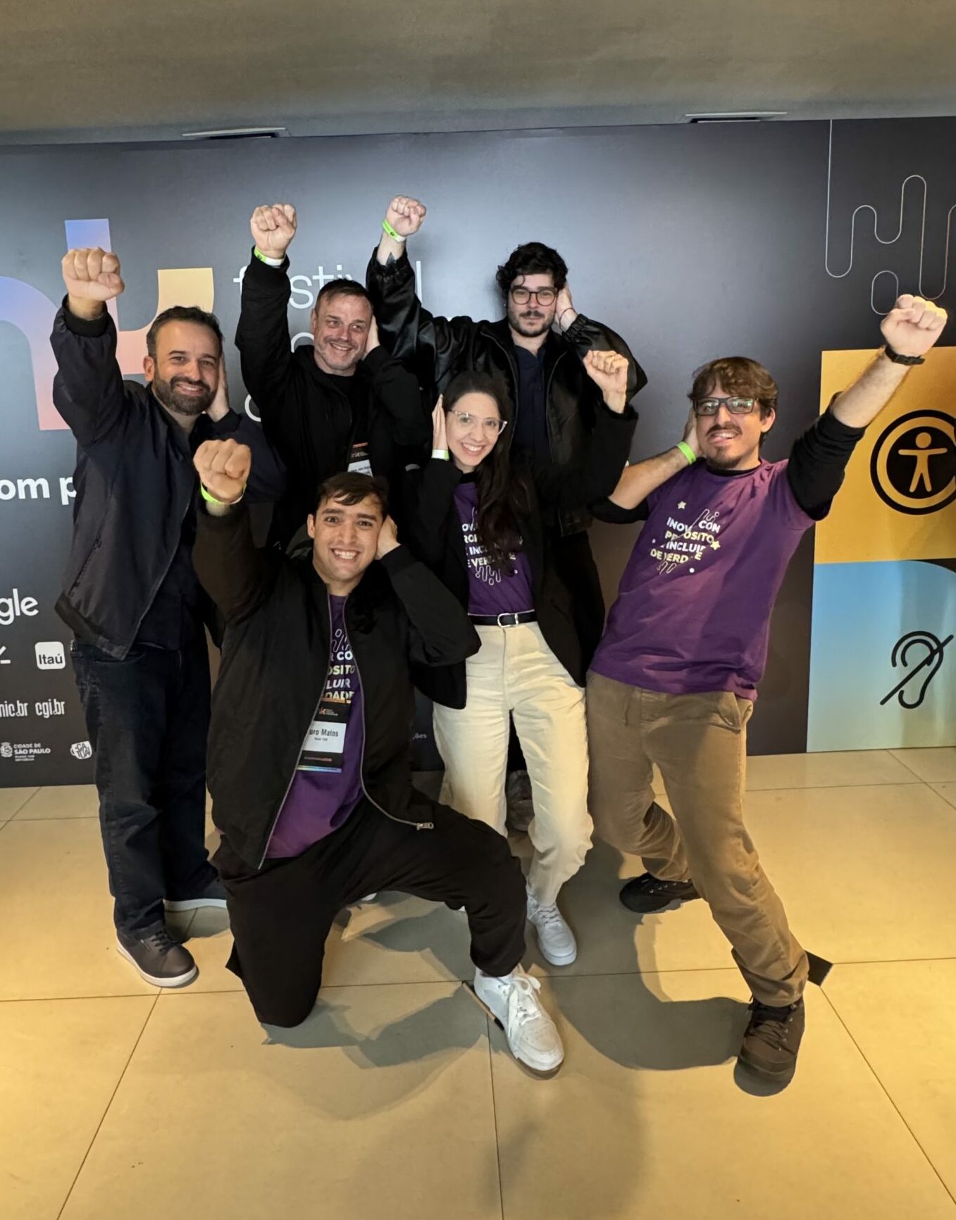 Six Deaf HandTalkers are smiling while striking the Deaf Power pose: one hand placed on their own ear and the other hand raised, connecting deafness with a position of power.